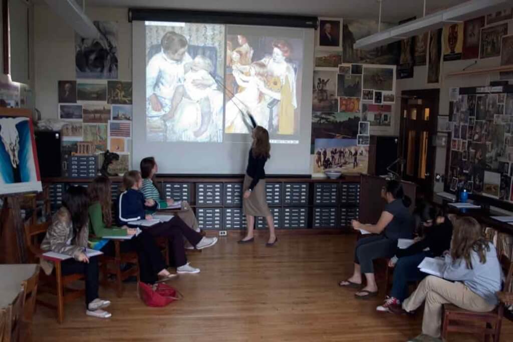 In a room with the appearance of studio, with many artworks and pictures on the walls and a file cabinet on the floor, a woman uses a point to show a detail in the slide of a painting being projected on a screen. On each side of the room a group of young people sitting on chairs looks at the image on the screen. The image shows two paintigs, each of a woman interacting with a child on their lap.
