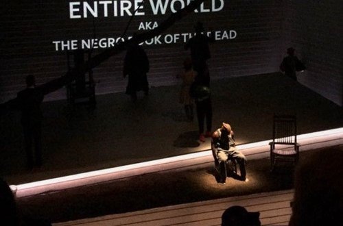 Photo by anonymous student photographer, depicting a mostly dark stage with a man, spotlighted, sitting on a chair, arms down, head down, next to an empty chair. The white, all-caps text above him reads -- partially obscured -- "black man in the whole entire world AKA the Negro Book of the Dead."